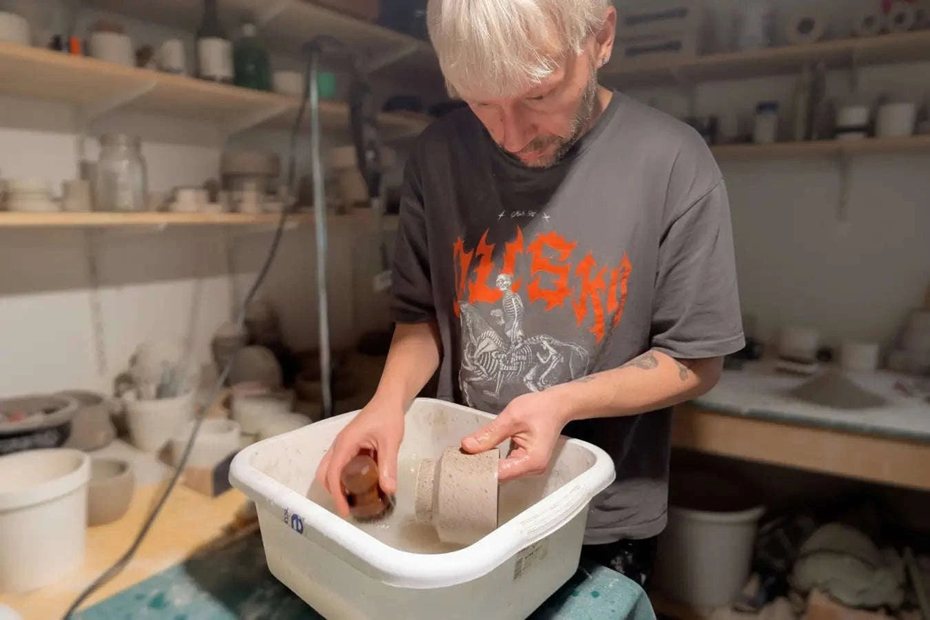Ein Handwerker mit kurzen hellen Haaren und einem grauen T-Shirt wäscht und schleift einen gegossenen Beton-Pflanztopf per Hand in einer weißen Kunststoffwanne. Im Hintergrund ist die authentische Werkstatt mit Regalen voller Formen und Werkzeuge zu sehen.