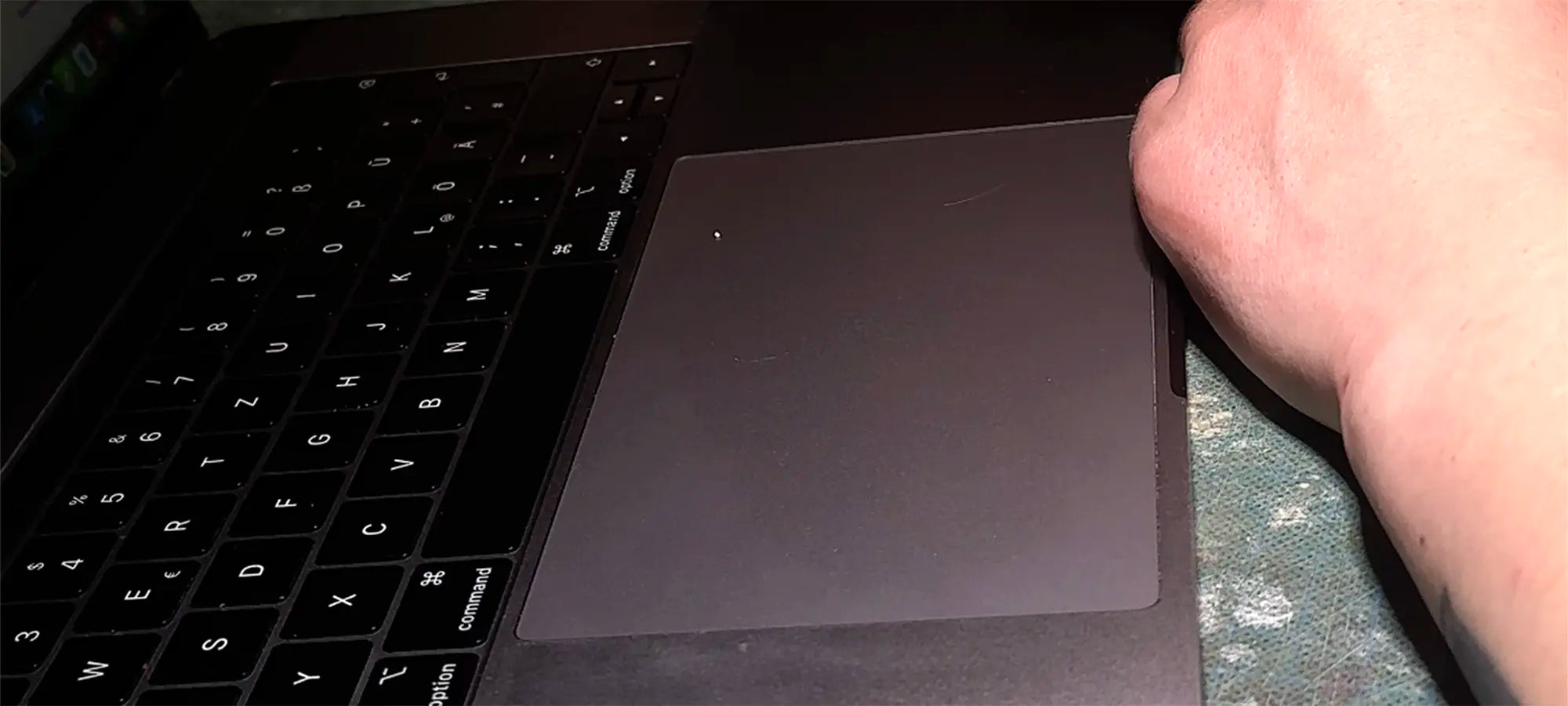 Winkelaufnahme eines dunklen Laptops (MacBook-Stil) mit gut sichtbarer Tastatur und Trackpad. Eine Hand ruht am rechten Gehäuserand.