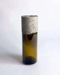Schmale Vase aus braunem Upcycling-Glas mit grober Betonmanschette vor hellem Hintergrund