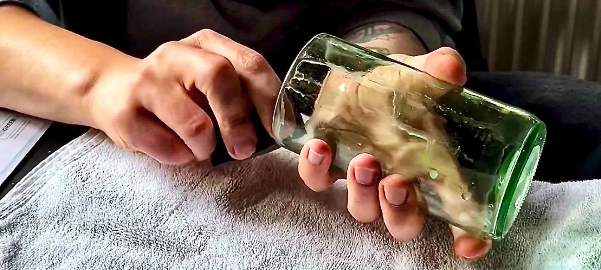 Nahaufnahme im Querformat: Zwei Hände bearbeiten sorgfältig die frisch geschliffene Kante eines upgecycelten, grünen Trinkglases aus einer Weinflasche. Das Glas liegt dabei stabil auf einem hellen Handtuch.