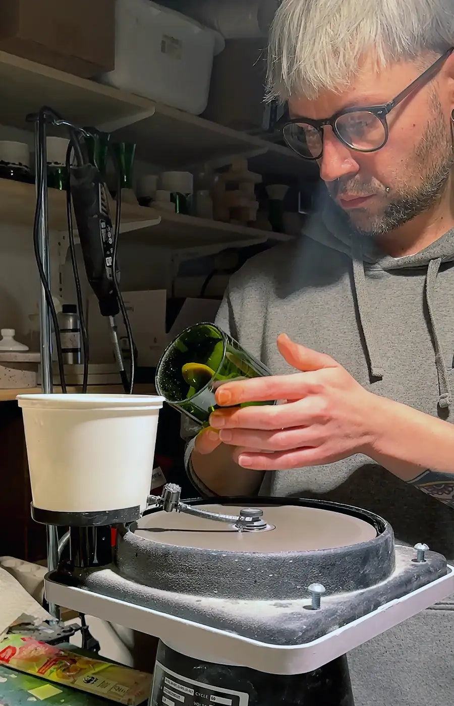 Ein Mann mit kurzen weißen Haaren und Brille, der einen grauen Kapuzenpullover trägt, arbeitet in einer Werkstatt. Er steht vor einer horizontalen Glasschleifmaschine mit Wasserkühlung und hält ein Stück dunkles, grünes Flaschenglas gegen die Schleifscheibe. Seine Hände sind präzise positioniert, und er blickt konzentriert auf seine Arbeit, was die handwerkliche Qualität und die handgeprüfte Verarbeitung von Trinkgläsern aus Weinflaschen zeigt.