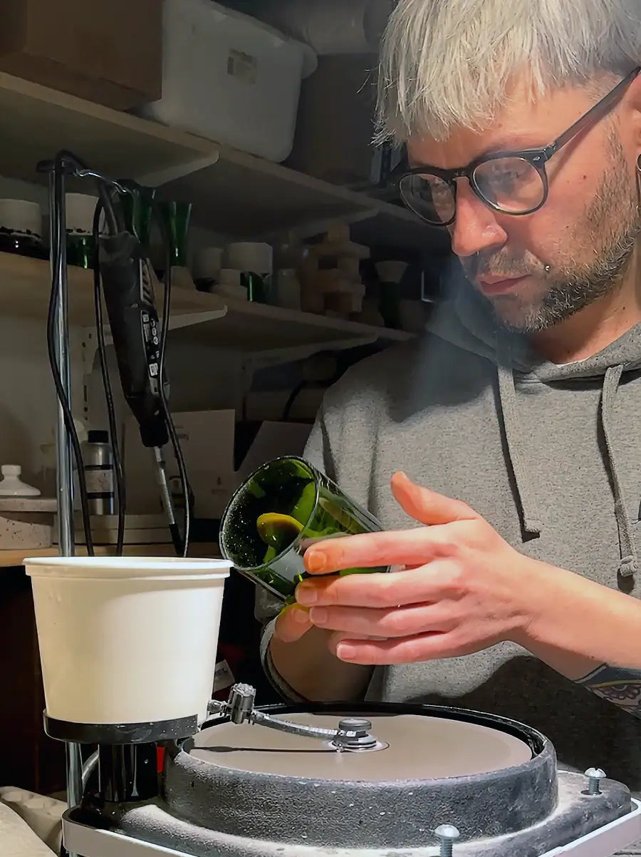 Ein Mann mit kurzen weißen Haaren und Brille, der einen grauen Kapuzenpullover trägt, arbeitet in einer Werkstatt. Er steht vor einer horizontalen Glasschleifmaschine mit Wasserkühlung und hält ein Stück dunkles, grünes Flaschenglas gegen die Schleifscheibe. Seine Hände sind präzise positioniert, und er blickt konzentriert auf seine Arbeit, was die handwerkliche Qualität und die handgeprüfte Verarbeitung von Trinkgläsern aus Weinflaschen zeigt.