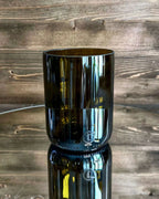 Trinkglas aus antikgrünem Upcycling-Glas auf Spiegelplatte vor dunkler Holzwand