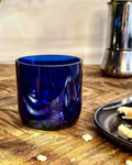 Seltenes Blaues Trinkglas aus Weinflasche auf Holztisch neben schwarzem Teller und Espressokocher