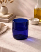 Blaues Trinkglas auf hellem Tisch mit Vase, Büchern und Schale im Hintergrund