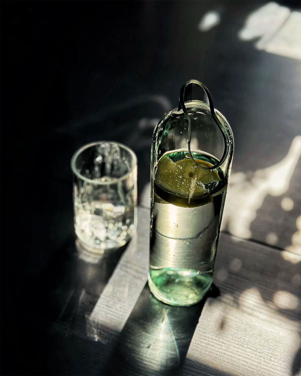 Upcycling-Wasserkaraffe aus einer Weinflasche mit Zitronenscheiben neben einem gefüllten Wasserglas auf dunkler Holzoberfläche mit Licht- und Schattenspiel