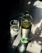 Upcycling-Wasserkaraffe aus einer Weinflasche mit Zitronenscheiben neben einem gefüllten Wasserglas auf dunkler Holzoberfläche mit Licht- und Schattenspiel