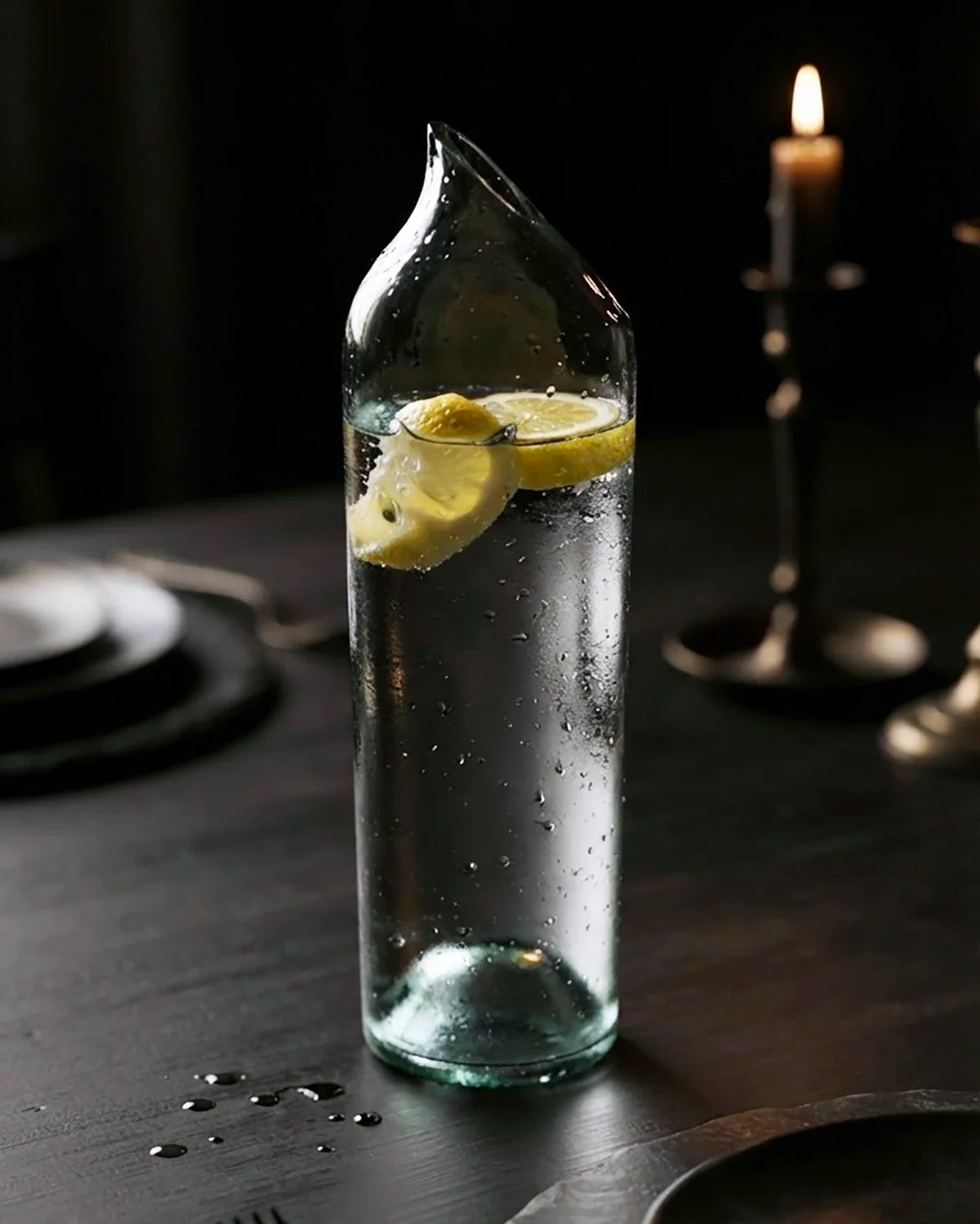 Glaskaraffe aus Upcycling-Altglas gefüllt mit Zitronenwasser auf einem gedeckten Esstisch neben einer brennenden Kerze
