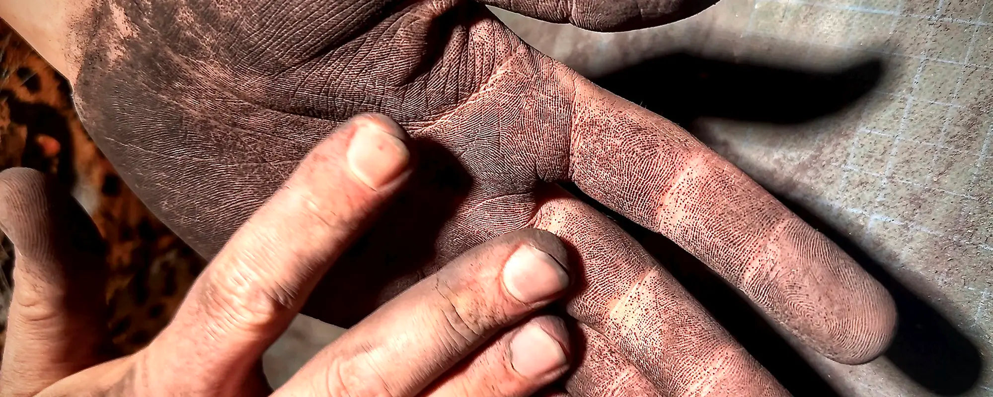Extreme Nahaufnahme im Querformat von zwei arbeitenden Händen: Eine geöffnete Handfläche und Finger sind komplett mit feinem, dunklem Zementstaub oder Betonpulver bedeckt, sodass die Hautstrukturen und Fingerabdrücke stark betont werden. Die andere Hand berührt sie sanft.