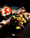 Zwei Antikgrüne Upcycling-Tapasschalen aus Glasflaschen, gefüllt mit Oliven sowie Tomate-Mozzarella-Salat, angerichtet auf einem Holzbrett mit frischem Brot bei starkem Licht und Schattenspiel.