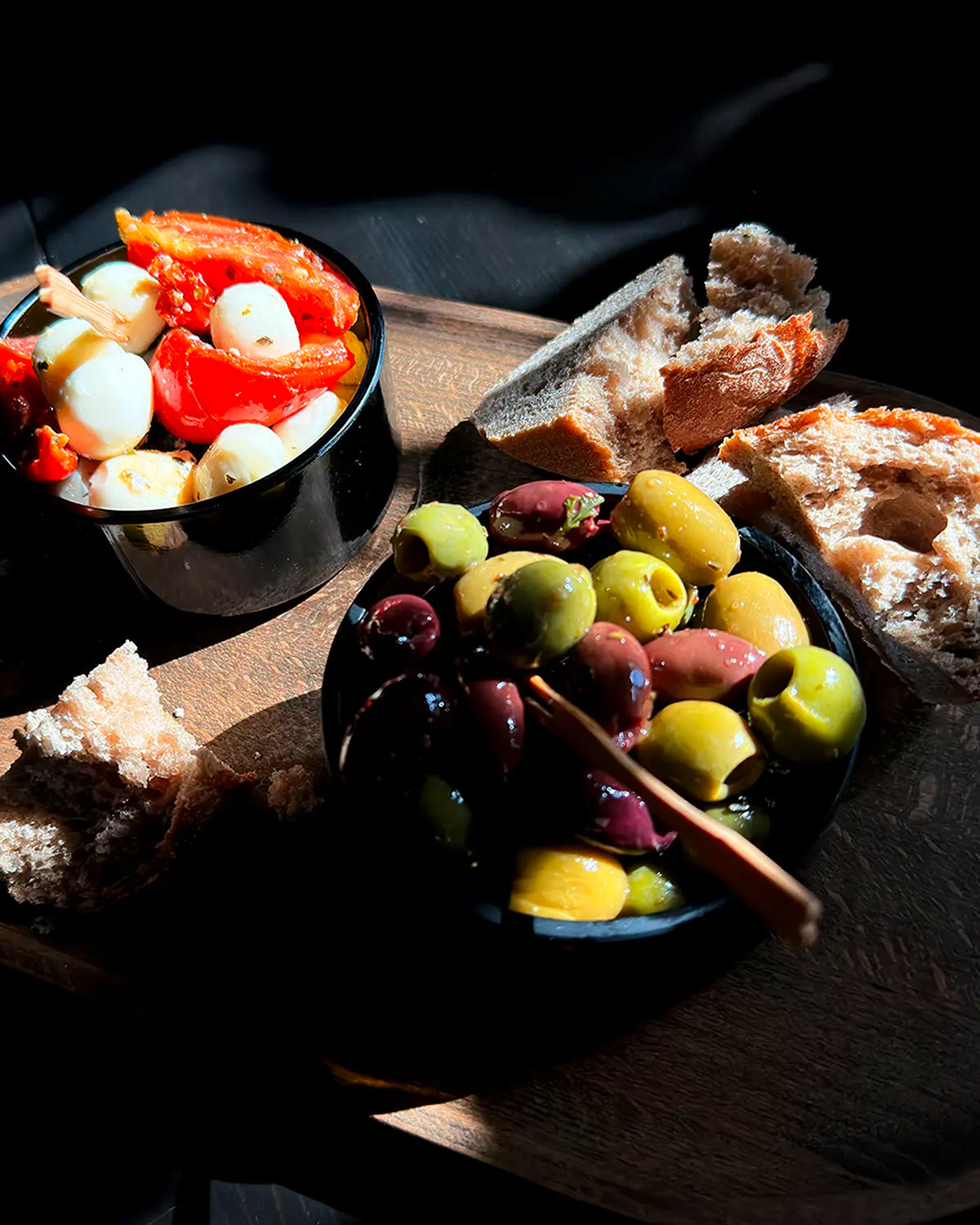 Zwei Antikgrüne Upcycling-Tapasschalen aus Glasflaschen, gefüllt mit Oliven sowie Tomate-Mozzarella-Salat, angerichtet auf einem Holzbrett mit frischem Brot bei starkem Licht und Schattenspiel.