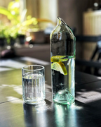 Frontalaufnahme einer Upcycling-Wasserkaraffe aus einer grünen Weinflasche mit Zitronenscheiben und einem Trinkglas auf einem Holztisch