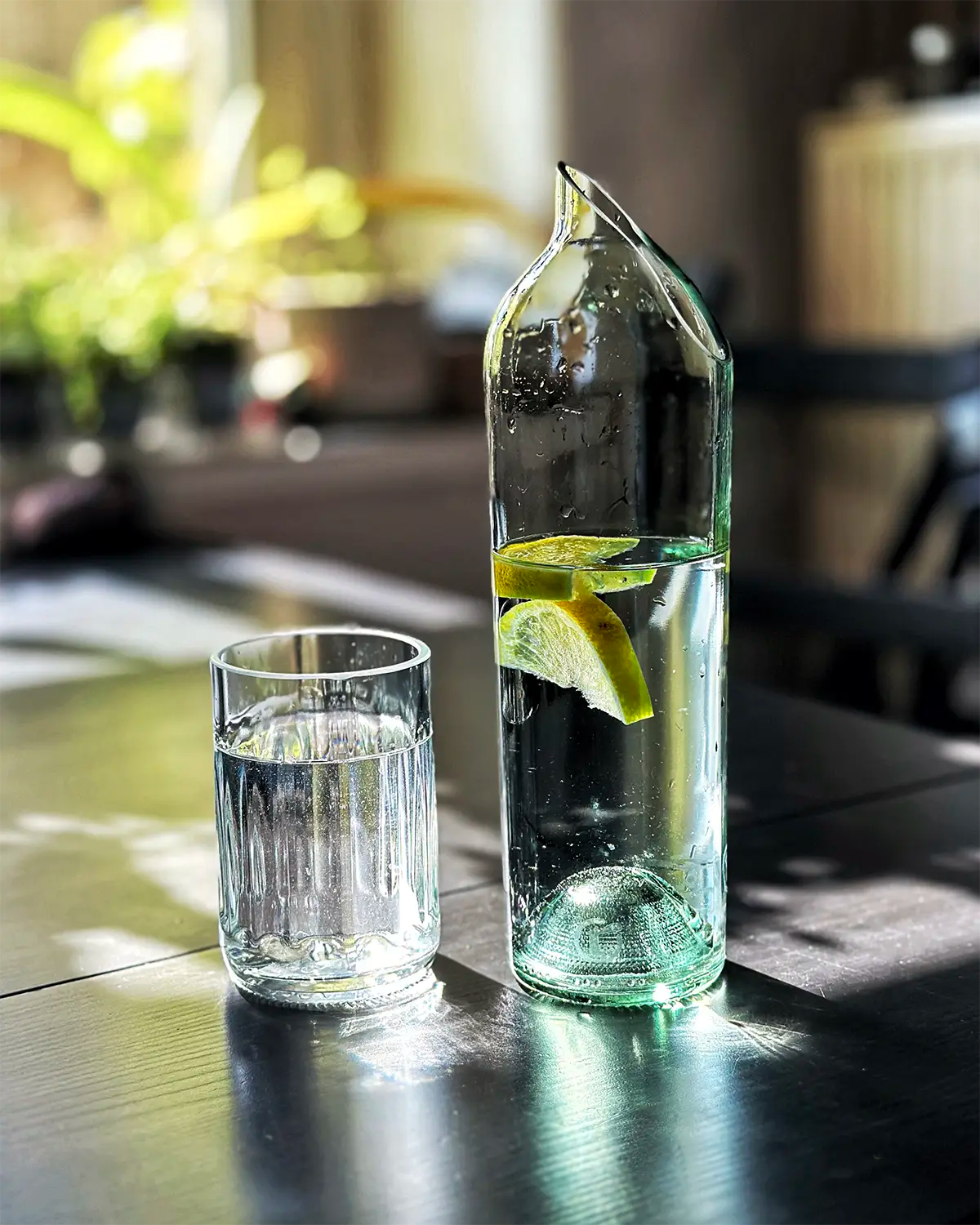 Frontalaufnahme einer Upcycling-Wasserkaraffe aus einer grünen Weinflasche mit Zitronenscheiben und einem Trinkglas auf einem Holztisch