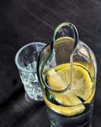 Nahaufnahme des geschliffenen, geschwungenen Ausgusses einer Glaskaraffe aus Altglas mit Zitronenscheibe und Trinkglas im Hintergrund