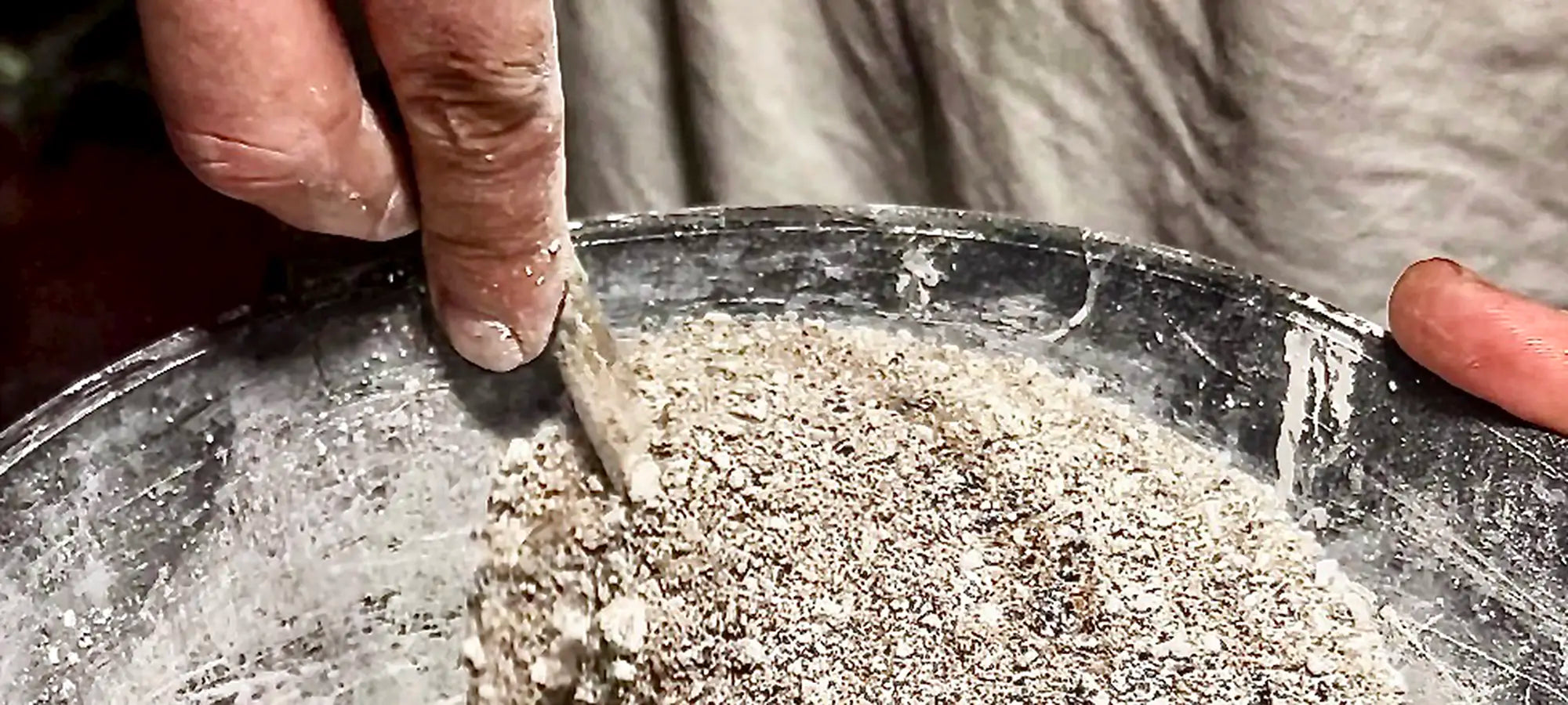 Extreme Makroaufnahme im Querformat: Eine Hand mischt mit einem Werkzeug eine feine, sandige Beton- und Zementmischung in einem dunklen, abgenutzten Baueimer. An den Fingern sind feine Materialspuren der Handarbeit zu sehen.